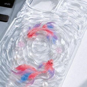 May include: Clear phone case featuring two colorful koi fish with red, pink, and blue hues, along with pearl and pink bead accents. The case has a water-like design, creating a unique and artistic look.