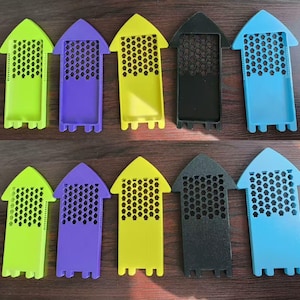 May include: A collection of colorful plastic beekeeping queen excluders. The excluders are in various colors, including green, purple, yellow, black, and blue. Each has a honeycomb-patterned section and an arrow-shaped top.