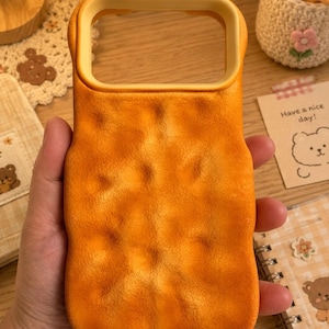 May include: A rectangular phone case with a textured, uneven surface resembling a golden-brown chicken nugget. The case features a smooth, raised yellow border around the camera cutout area, providing a distinct contrast to the bumpy, orange-toned main body of the protective cover.