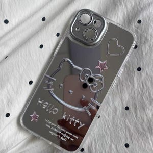 May include: A clear phone case with a mirrored back featuring a Hello Kitty design. The case has a silver Hello Kitty graphic with a pink bow and stars. The text "Hello Kitty" is printed on the case, along with the quote "May you become your own sun without needing anyone's light."