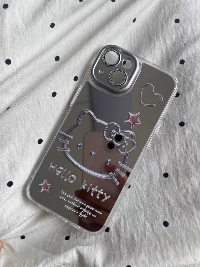 May include: A clear phone case with a mirrored back featuring a Hello Kitty design. The case has a silver Hello Kitty graphic with a pink bow and stars. The text "Hello Kitty" is printed on the case, along with the quote "May you become your own sun without needing anyone's light."