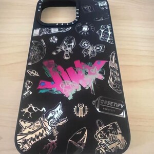 Peut inclure: Coque de t&eacute;l&eacute;phone noire avec un design graphique argent&eacute; et rose. Le design pr&eacute;sente un requin, un papillon et le mot "JUNK" en lettres roses et blanches. La coque porte &eacute;galement le texte "CASETIFY" en lettres blanches.
