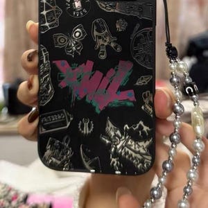 Peut inclure: Coque de t&eacute;l&eacute;phone noire avec un design graphique color&eacute; et un cache objectif d'appareil photo argent&eacute; avec des strass. La coque est munie d'une dragonne de t&eacute;l&eacute;phone &agrave; perles noires et blanches.