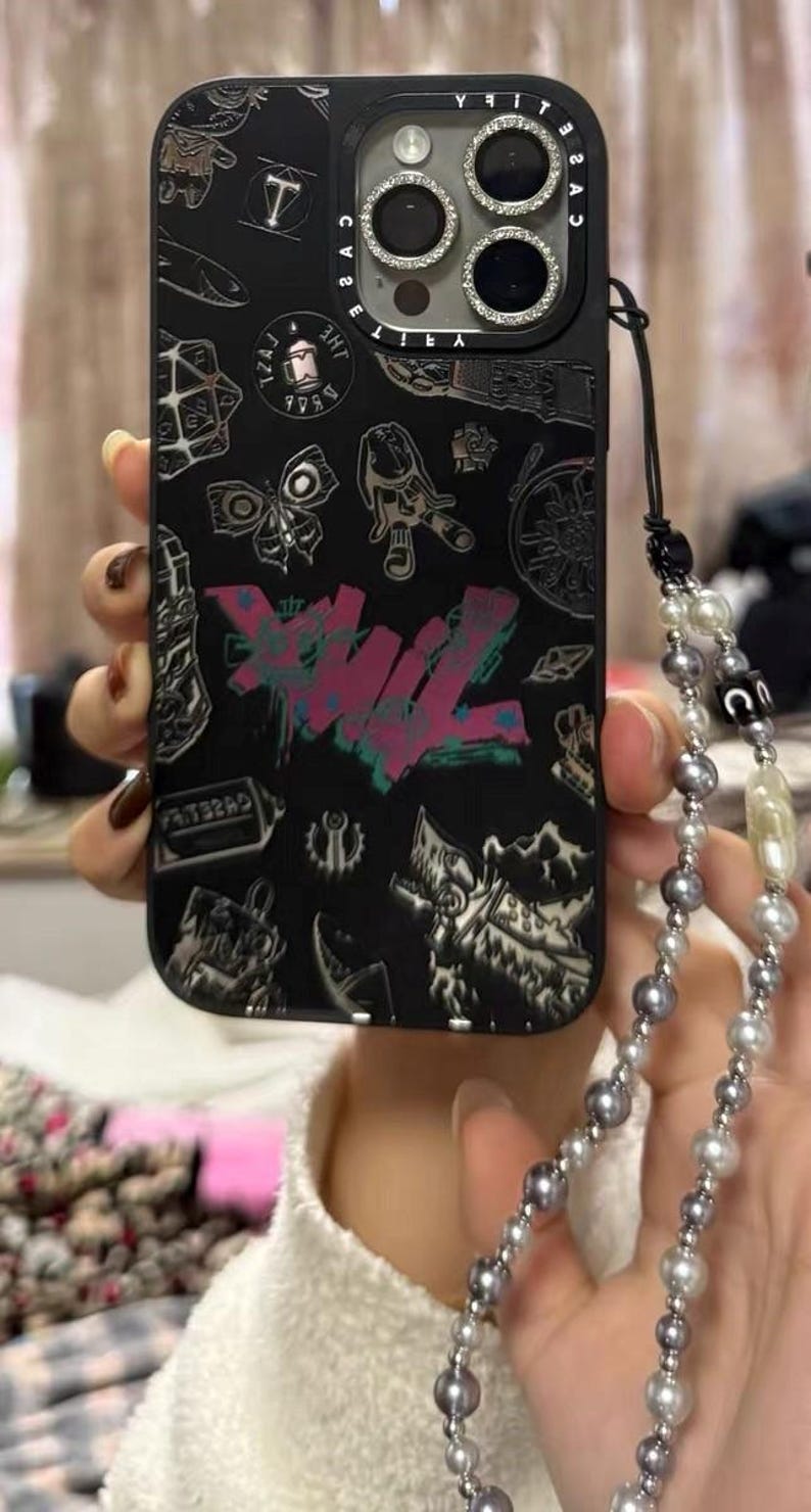 Peut inclure: Coque de t&eacute;l&eacute;phone noire avec un design graphique color&eacute; et un cache objectif d'appareil photo argent&eacute; avec des strass. La coque est munie d'une dragonne de t&eacute;l&eacute;phone &agrave; perles noires et blanches.