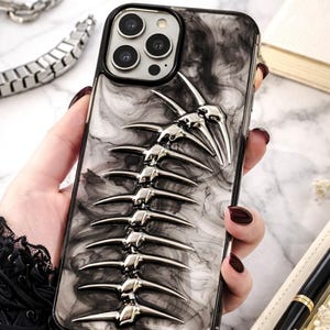 May include: A black and gray phone case with a silver spine design. The case has a marbled pattern and a raised, metallic spine detail. The phone case is held in a hand with dark red nail polish.