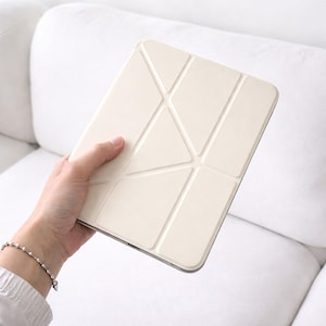 Puede incluir: Una funda de tableta de color crema con un diseño geométrico. La funda, sostenida en una mano, presenta una serie de líneas que se cruzan formando triángulos y rectángulos. Tiene un acabado liso y mate.