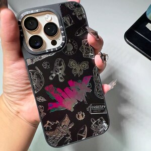 Peut inclure: Une coque de t&eacute;l&eacute;phone iPhone noire avec un design argent&eacute; et noir. La coque pr&eacute;sente une vari&eacute;t&eacute; de graphismes, notamment un papillon, une bombe de peinture et le mot "CASEIFY".