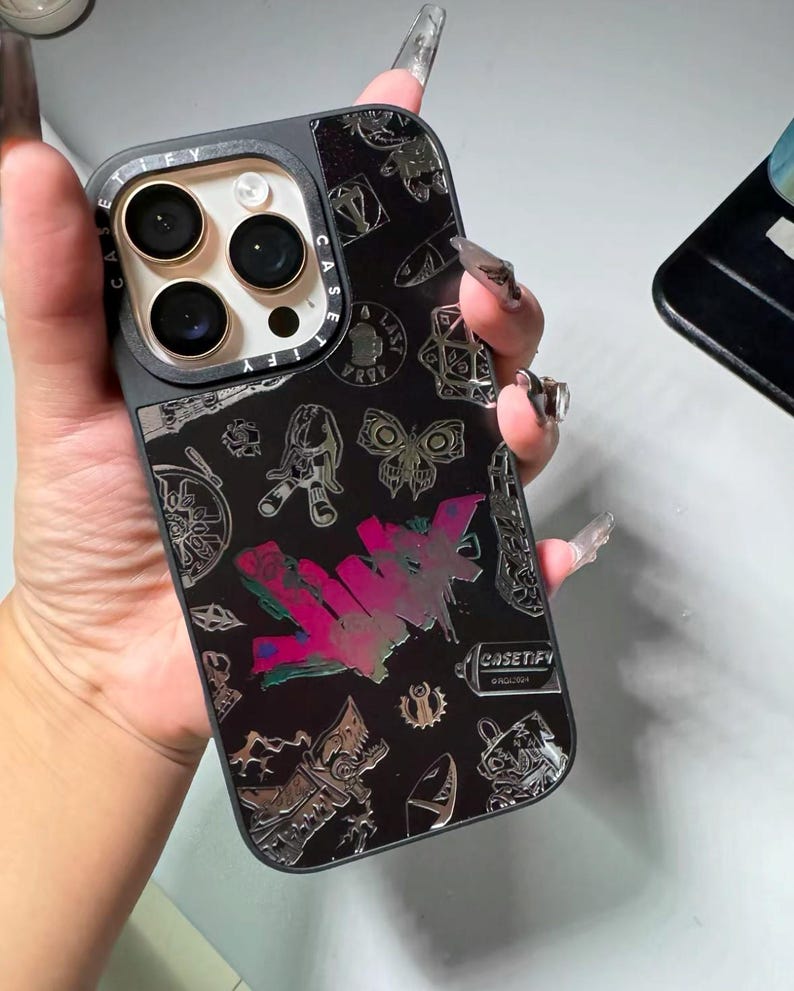 Peut inclure: Une coque de t&eacute;l&eacute;phone iPhone noire avec un design argent&eacute; et noir. La coque pr&eacute;sente une vari&eacute;t&eacute; de graphismes, notamment un papillon, une bombe de peinture et le mot "CASEIFY".