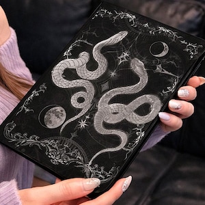 May include: Black notebook with a silver snake design. The cover features two intertwined snakes, moons, and stars. The notebook has an ornate border and is held in a person's hands.