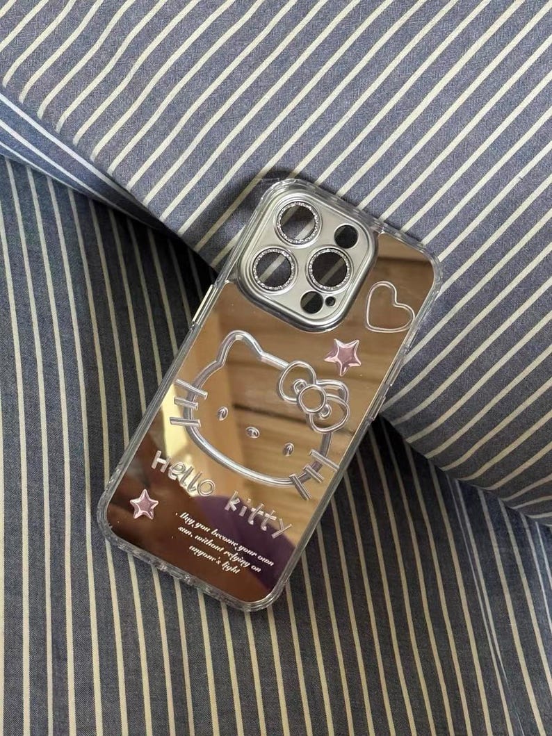 May include: A silver mirror phone case with a Hello Kitty design. The case has a pink heart and stars. The text on the case reads "Hello Kitty. Way you become your own ann, without refying on anyone's light."