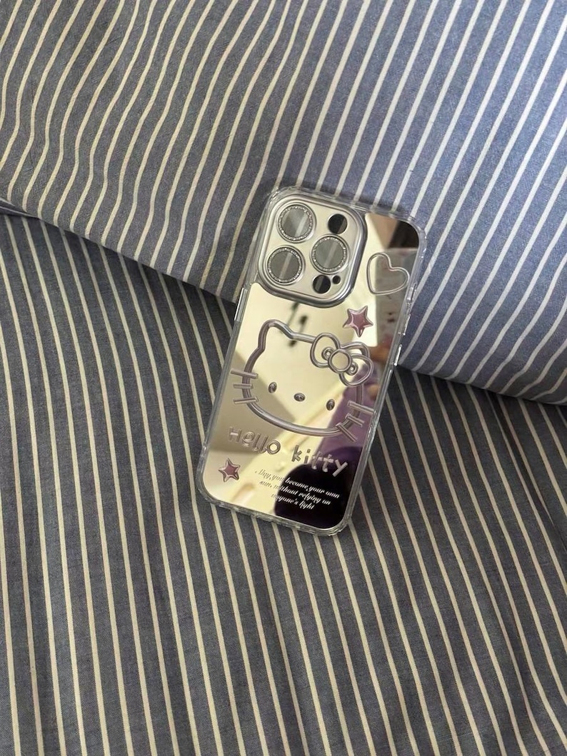 May include: A clear phone case with a mirrored back featuring a Hello Kitty design. The case has a cutout for the camera lens and a pink heart on the back. The text "Hello Kitty" is printed on the case, along with the phrase "Because your smile is the most beautiful thing in the world."