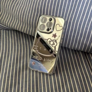 May include: A silver phone case with a Hello Kitty design. The case has a clear back and a silver frame. The Hello Kitty design is in the center of the case and is surrounded by a pink heart and two gold stars.