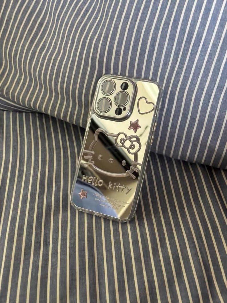 May include: A silver phone case with a Hello Kitty design. The case has a clear back and a silver frame. The Hello Kitty design is in the center of the case and is surrounded by a pink heart and two gold stars.