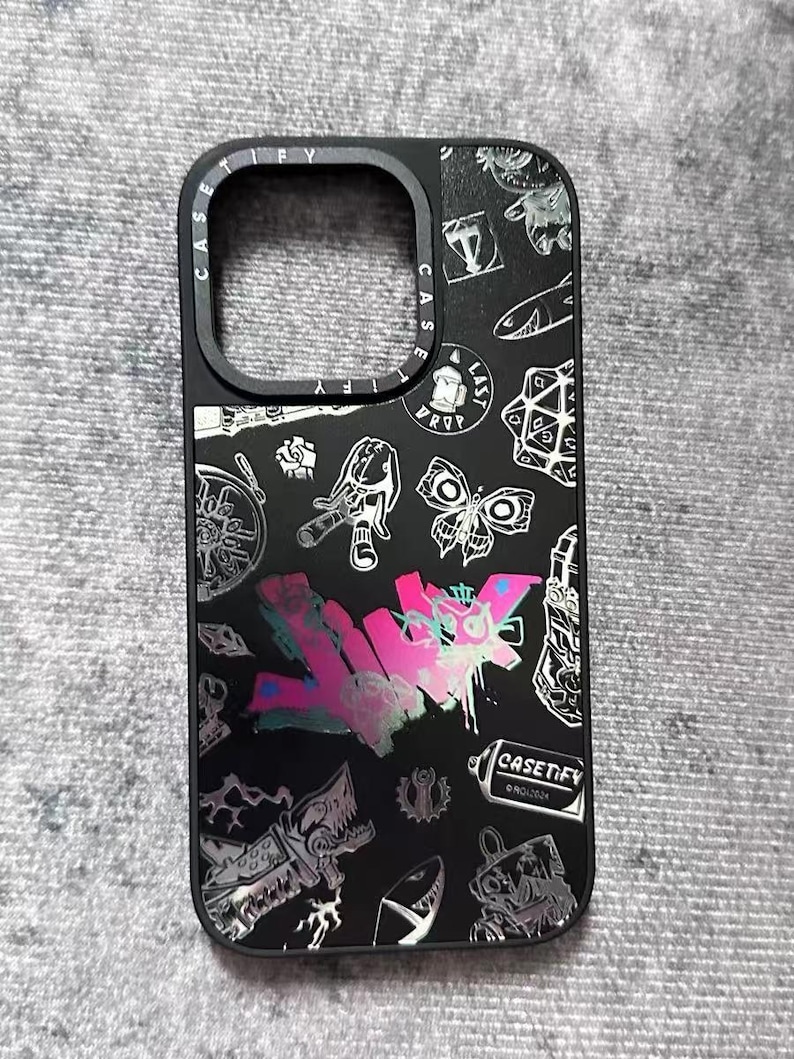 Peut inclure: Coque de t&eacute;l&eacute;phone noire avec un design graphique argent&eacute; et rose repr&eacute;sentant un papillon, un cr&acirc;ne, une bombe de peinture et le texte "CASETIFY".