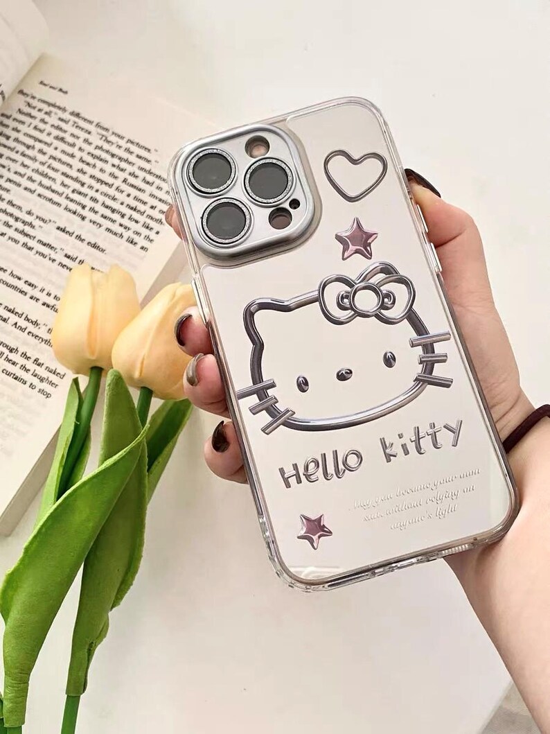 May include: Clear phone case with a Hello Kitty design. The case has a silver outline of Hello Kitty with a pink bow and a pink star. The case also has a silver heart and two silver stars.