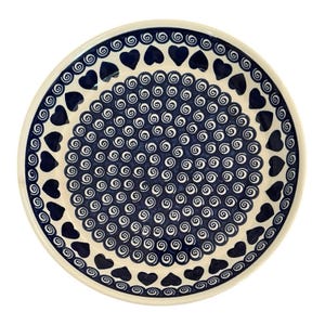 May include: A round ceramic plate featuring a cream-colored base with a dark blue pattern. The center is filled with small white spirals, surrounded by a border of dark blue hearts and a final outer rim of small white spirals on a blue band.