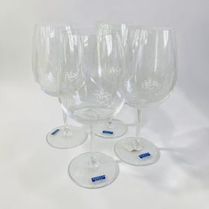 May include: A set of four clear glass wine glasses with long stems and rounded bowls. Each glass features a small blue and white rectangular sticker on the base or stem that reads Marquis by Waterford, indicating the brand of the glassware.
