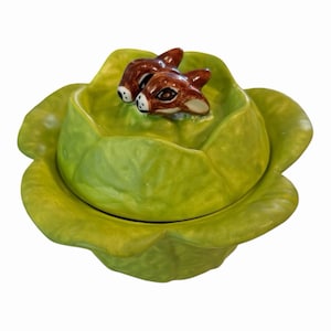 May include: A ceramic cabbage-shaped dish with a lid, featuring two brown and white deer figurines. The dish is a vibrant green color, with a glossy finish. The deer are detailed with brown fur and white accents.