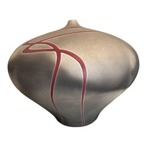 May include: A rounded ceramic vase with a metallic silver finish and a narrow opening at the top. The surface features a series of deep red, curved lines that wrap around the body of the vessel, creating a distinct contrast against the smooth, reflective gray texture of the exterior.