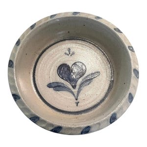 May include: A round ceramic bowl with a speckled off-white glaze. The interior features a hand-painted blue heart-shaped floral motif at the center. The thick, textured rim is decorated with a repeating pattern of small, dark blue painted leaf shapes.
