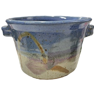 May include: A handmade ceramic bowl with a blue exterior and a speckled, textured surface. The bowl features two small handles and a painted design with shades of purple, tan, and white. This unique piece is perfect for serving or display.
