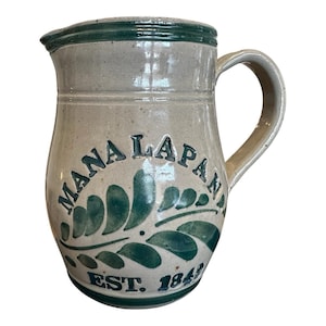 May include: A beige ceramic pitcher with a dark green rim and base. The front features the dark green text MANA LAPAN above a painted green leaf design and the text EST. 1843. It has a curved handle on the right side and a pouring spout.