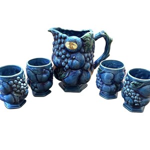 May include: A ceramic pitcher and four matching cups in a deep blue glaze. The set features a raised, three-dimensional relief of grapes and various fruits. The pitcher includes a handle and a gold-colored label with the text "SARREGUEMINES FRANCE".