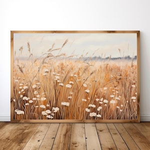 Puede incluir: Una pintura de paisaje enmarcada representa un campo de hierbas altas y doradas y flores silvestres blancas. La obra de arte tiene una paleta de colores cálidos con un cielo azul claro y un marco de madera. La pintura se exhibe sobre un suelo de madera.