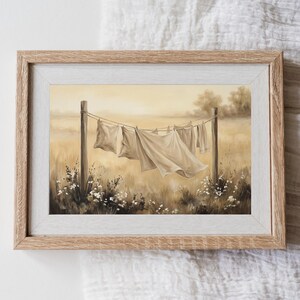 May include: A framed painting depicting white linens hanging on a clothesline between two wooden posts in a field. The scene features a soft, warm, sepia-toned color palette with blurred trees in the background and small white flowers in the foreground.