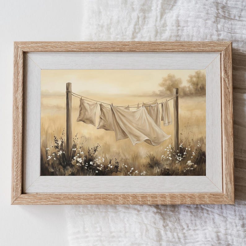 May include: A framed painting depicting white linens hanging on a clothesline between two wooden posts in a field. The scene features a soft, warm, sepia-toned color palette with blurred trees in the background and small white flowers in the foreground.