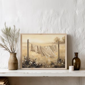 May include: A rectangular framed print featuring a painting of white linens hanging on a clothesline between two wooden posts in a field. The scene is rendered in soft, warm, sepia-toned colors with visible wildflowers at the base of the posts.