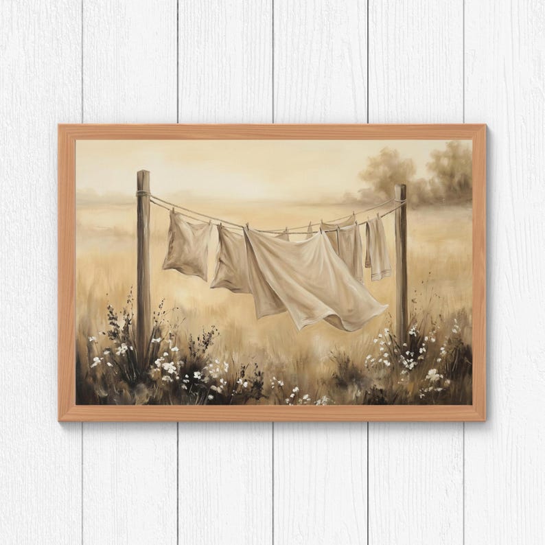 May include: A framed landscape painting in sepia tones depicting white laundry hanging on a clothesline between two wooden posts. The scene features a field of tall grass and small white flowers in the foreground, with a soft, hazy horizon.