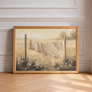 May include: A framed landscape painting in a light wood frame depicting white laundry hanging on a clothesline between two wooden posts in a field. The scene features soft beige and cream tones with small white flowers at the base of the posts.