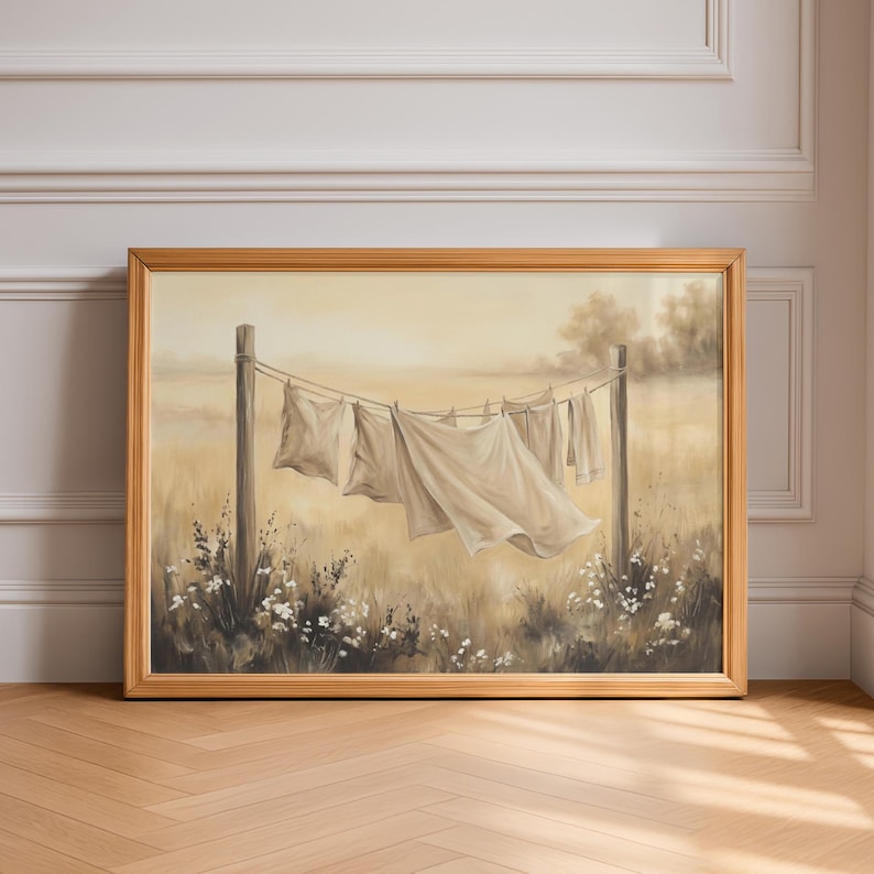 May include: A framed landscape painting in a light wood frame depicting white laundry hanging on a clothesline between two wooden posts in a field. The scene features soft beige and cream tones with small white flowers at the base of the posts.