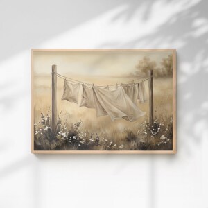May include: A framed landscape painting depicting white linens hanging on a clothesline between two wooden posts in a field. The scene features soft beige and brown tones with wildflowers at the base, capturing a serene outdoor setting.
