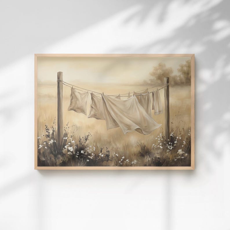 May include: A framed landscape painting depicting white linens hanging on a clothesline between two wooden posts in a field. The scene features soft beige and brown tones with wildflowers at the base, capturing a serene outdoor setting.