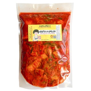 May include: A clear plastic bag filled with red and white Napa kimchi, a Korean fermented cabbage dish. The bag has a yellow label with the text "Seoulmate Fermented & Delicious" and "Napa Kimchi Made by a Real Korean Mama".