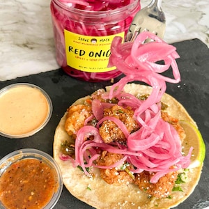 May include: A close-up of a taco filled with fried shrimp, pickled red onions, and coriander. The taco is on a black surface with a small container of salsa and a jar of pickled red onions. The jar is labelled "Seoul Mate Red Onion Sweet + Sour Pickles".