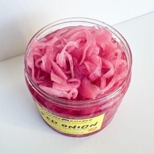 May include: A clear glass jar filled with bright pink pickled red onions. The jar has a yellow label that reads "Mama Kitchen" and "Red Onion" with a small graphic of a pickle.