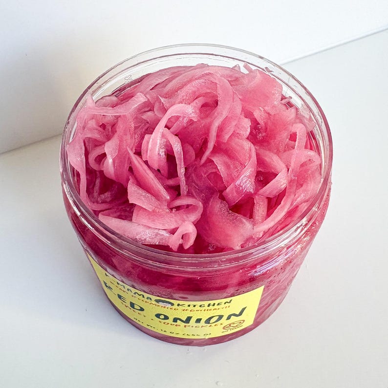 May include: A clear glass jar filled with bright pink pickled red onions. The jar has a yellow label that reads "Mama Kitchen" and "Red Onion" with a small graphic of a pickle.