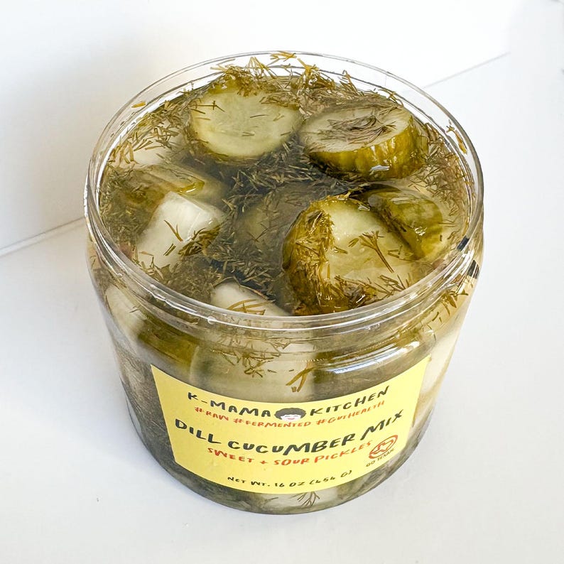 May include: A clear glass jar filled with dill pickle slices and dill. The jar has a yellow label that reads "K-Mama Kitchen Raw Fermented Goodness Dill Cucumber Mix Sweet + Sour Pickles Net Wt. 16 oz (454 g)"