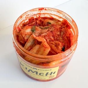 May include: A clear glass jar filled with red kimchi, a fermented Korean cabbage dish. The jar has a yellow label with the text "Mamao Kitchen Kimchi Authentic Korean Mama" and "Net Wt. 16 oz (454 g)".