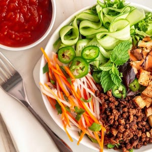 May include: A white bowl filled with a colourful salad with ingredients like shredded carrots, cucumbers, jalapenos, and ground meat. The salad is topped with fresh herbs and a side of red sauce.