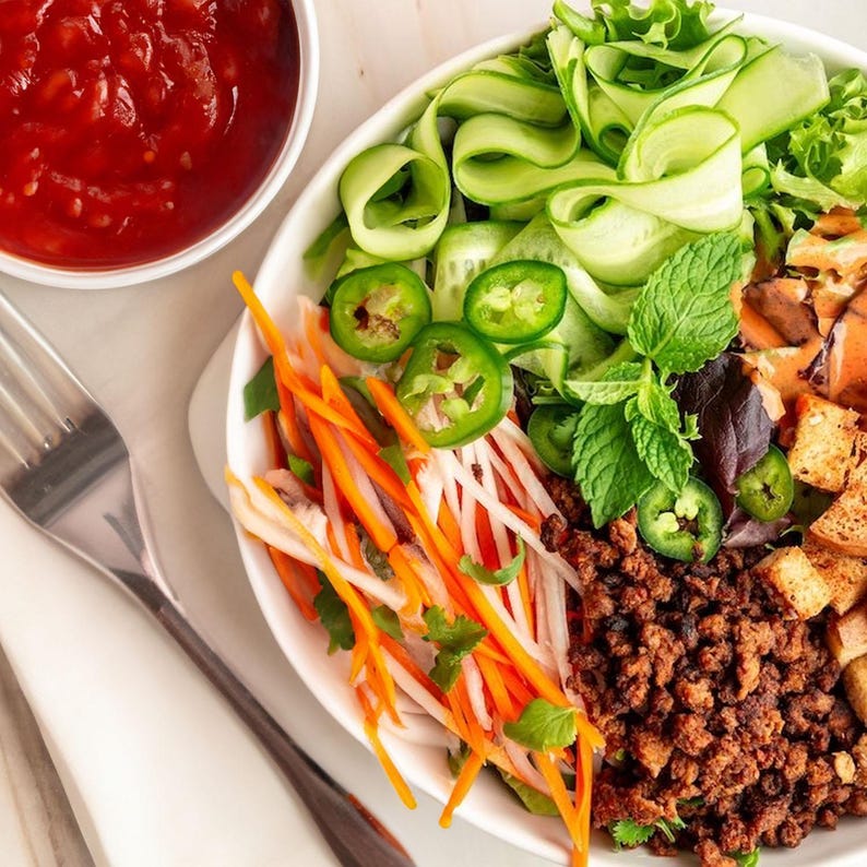 May include: A white bowl filled with a colourful salad with ingredients like shredded carrots, cucumbers, jalapenos, and ground meat. The salad is topped with fresh herbs and a side of red sauce.