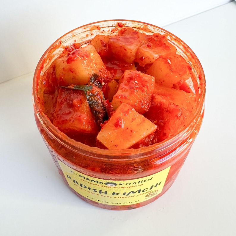 May include: A clear glass jar filled with red radish kimchi, a traditional Korean fermented food. The jar has a yellow label with the text "Mama Kitchen" and "Radish Kimchi" in black.