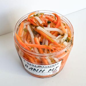 May include: A clear glass jar filled with a colourful mix of pickled carrots, daikon radish, and coriander. The jar has a white label with the text "K-MAMA KITCHEN REAL FERMENTED & GUT HEALTH BANH MI  viet soup style pickling net wt. 16 oz (454 g)"
