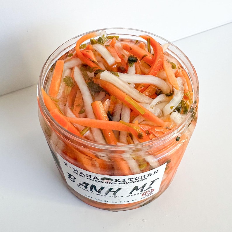 May include: A clear glass jar filled with a colourful mix of pickled carrots, daikon radish, and coriander. The jar has a white label with the text "K-MAMA KITCHEN REAL FERMENTED & GUT HEALTH BANH MI  viet soup style pickling net wt. 16 oz (454 g)"
