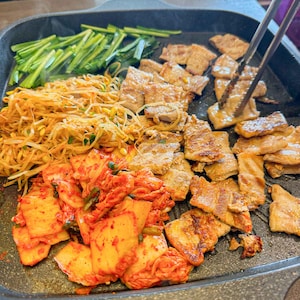 May include: A black cast iron pan filled with a variety of Korean BBQ ingredients, including thinly sliced pork belly, bean sprouts, kimchi, and spring onions.