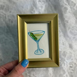 Puede incluir: Una pintura de acuarela de una copa de martini con tres aceitunas verdes. La pintura está enmarcada en un marco dorado.