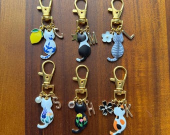 Puede incluir: Seis llaveros con forma de gato con cierres dorados. Cada gato tiene un diseño y un encanto únicos, que incluyen un limón, una estrella, una flor, una perla, una letra y detalles florales. Los gatos están pintados en varios colores, como blanco, negro y naranja.